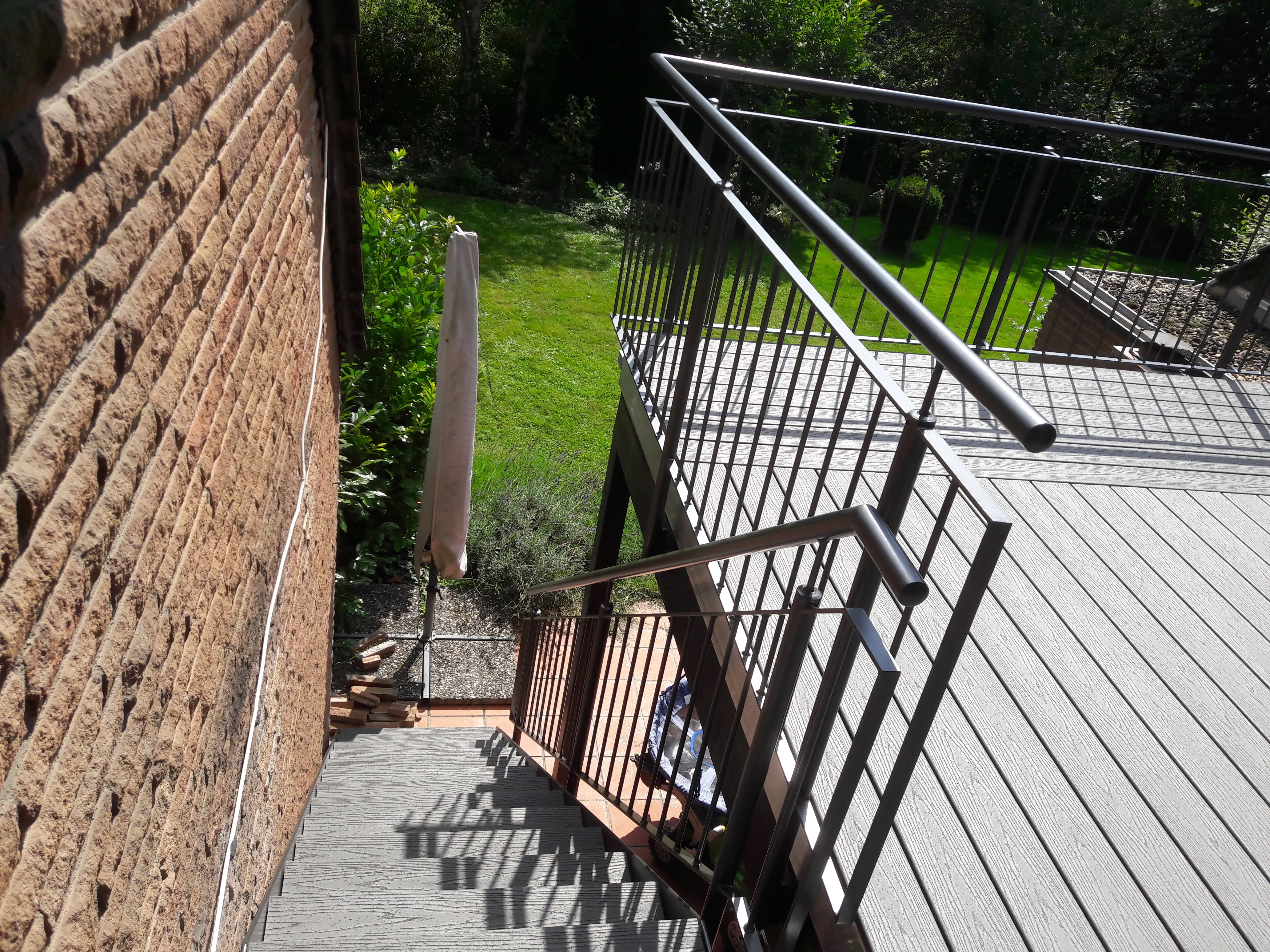 Blick von oben auf eine Außentreppe mit Metallgeländer neben einer Backsteinwand und einem geschlossenen Sonnenschirm auf einer Rasenfläche