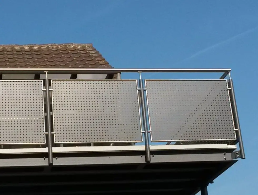 Balkon mit perforierten Metallplatten als Brüstung vor blauem Himmel und Teil eines Ziegeldachhauses