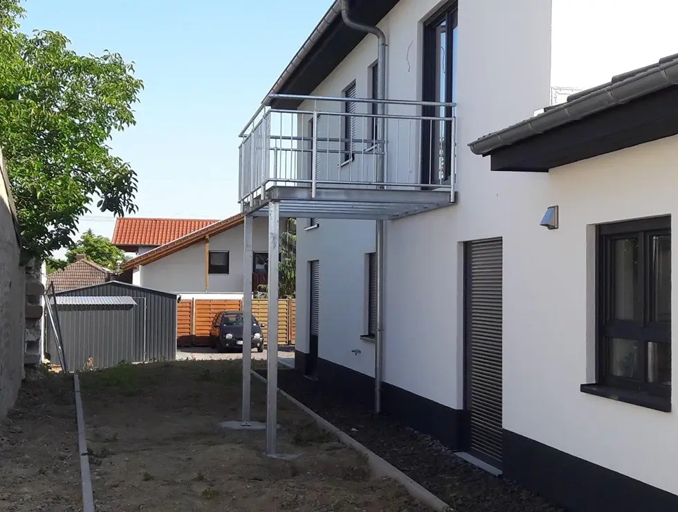 Moderner Balkon mit Metallgeländer an einem weißen zweistöckigen Haus mit dunklen Fensterläden und Tür