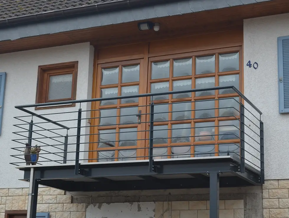Balkon mit schwarzem Metallgeländer vor zwei Holztüren mit Glasfenstern an einem hellen Haus mit Hausnummer 40