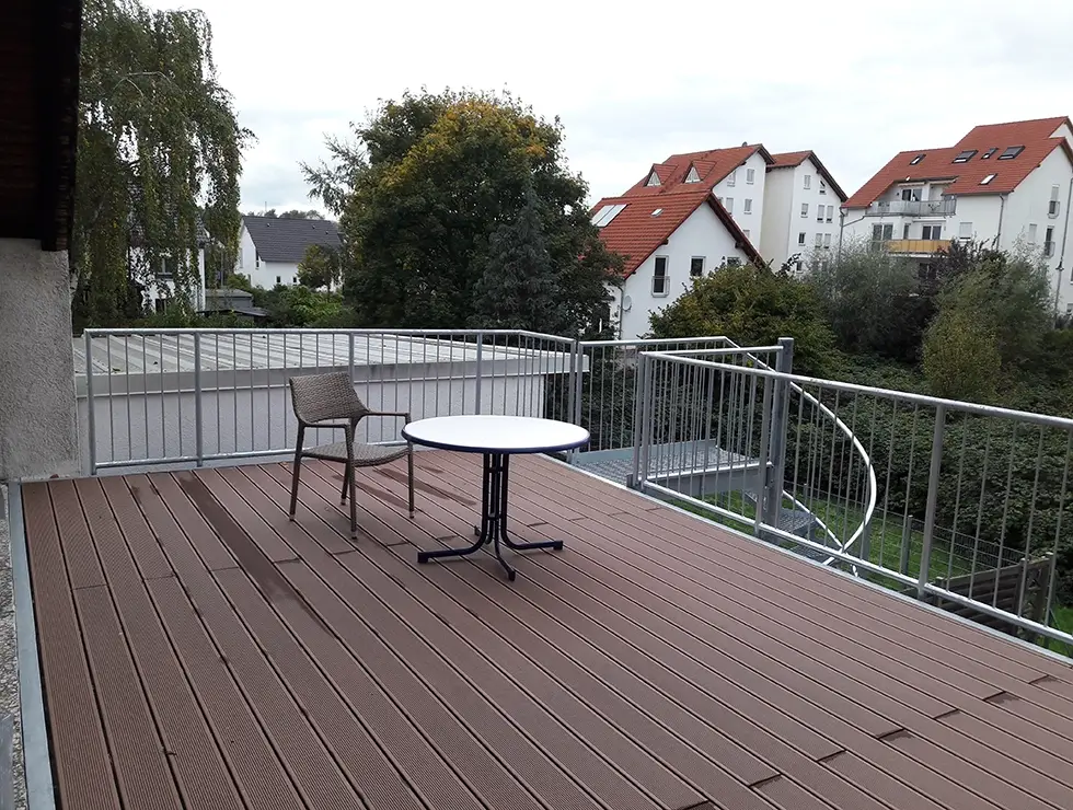 Balkon mit braunem Holzboden, einem runden weißen Tisch und einem Stuhl, umgeben von einem Metallgeländer mit einer Wendeltreppe