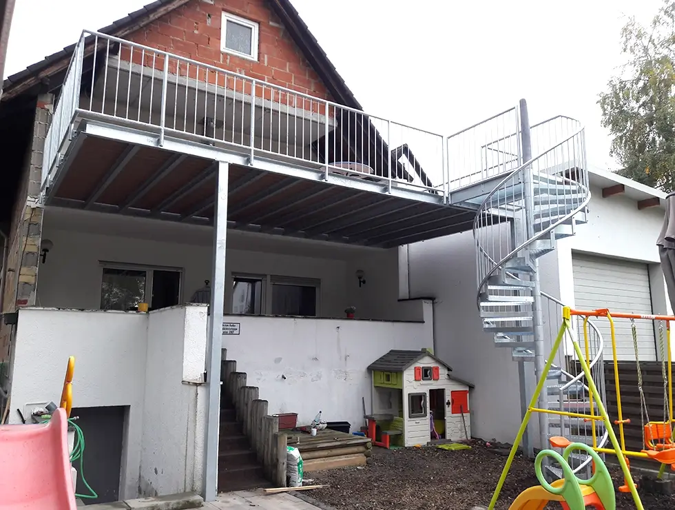 Außenbereich mit einer Metall-Spindeltreppe, die zu einer erhöhten Plattform an einem Haus führt, darunter Spielhaus und Kinderschaukel