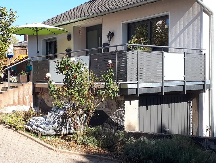 Einfamilienhaus mit Terrasse, die mit einem Metallgeländer mit blickdichten weißen Elementen eingefasst ist, davor ein Rosenstrauch und ein gepflasterter Weg
