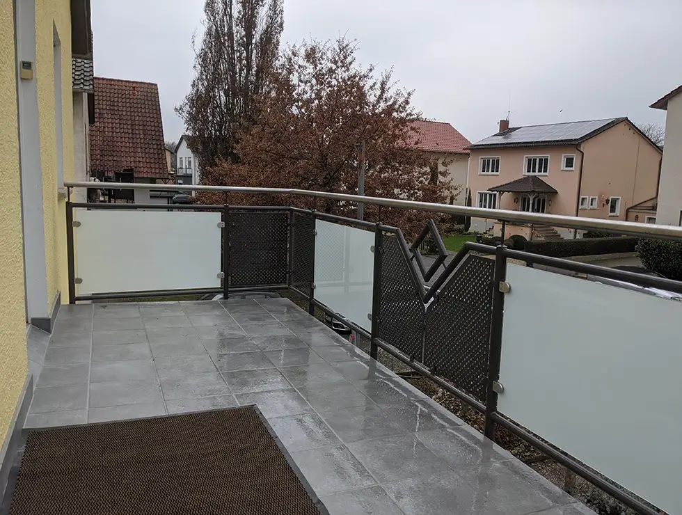 Balkon mit grauen Fliesen, braunem Teppich und Geländer mit Milchglas- und Metallfüllungen