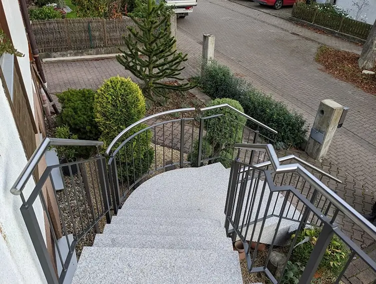 Außentreppe mit grauen Stufen und grauem Metallgeländer, umgeben von kleinen Büschen und einem Parkplatz im Hintergrund