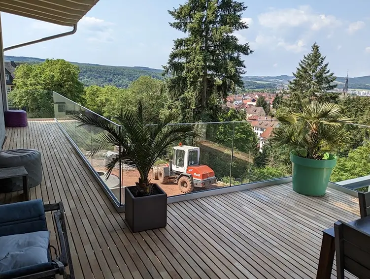 Terrasse mit Holzbohlen, Glasgeländer, zwei großen Pflanzen in Töpfen und Blick auf eine Stadt mit Bergen im Hintergrund