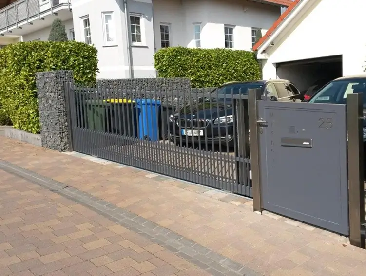 Schwarzer Metallzaun mit integriertem Briefkasten neben einem Holzgitterzaun vor einem Haus mit Hecke