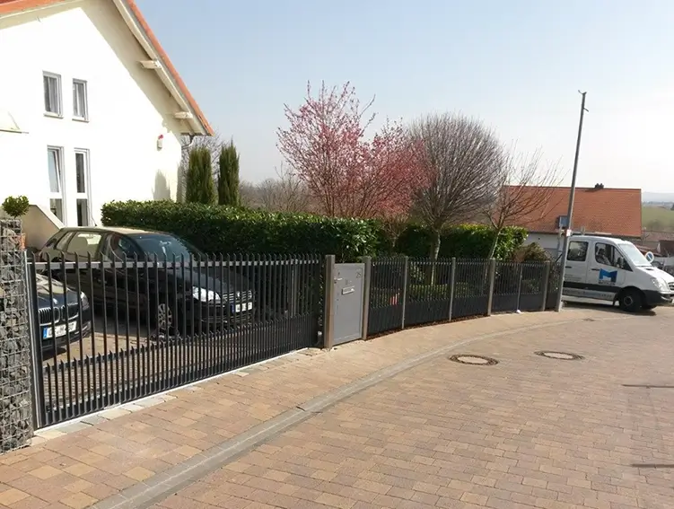 Schwarzer Metallzaun entlang eines Gehwegs mit Büschen und Bäumen dahinter sowie einem weißen Lieferwagen am Straßenrand