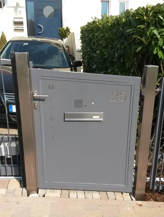 Graue Haustür mit Briefkasten, Gegensprechanlage und der Hausnummer 25 neben einem schwarzen Auto und grünen Hecken