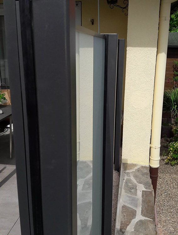 Mehrere vertikale, schwarze Metallrahmen mit Glasfüllung vor einer hellen Hauswand und einem Steinboden