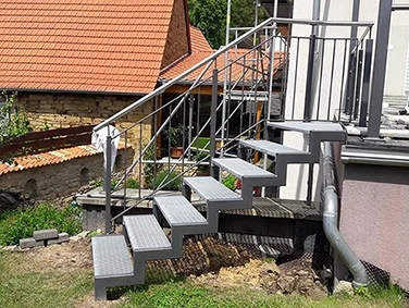 Außenmetalltreppe mit Gitterroststufen und Geländer, die an einem Haus ansetzt und über eine kleine Böschung führt