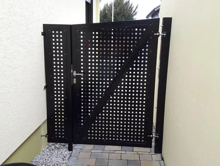 Schwarzes Metalltor mit quadratischen Aussparungen zwischen zwei hellen Wänden auf gepflastertem Boden