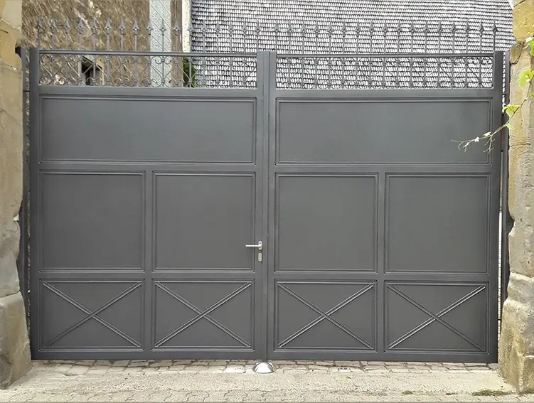 Großes, geschlossenes Tor aus Metall mit rechteckigen und diagonalen Mustern zwischen zwei Steinmauern