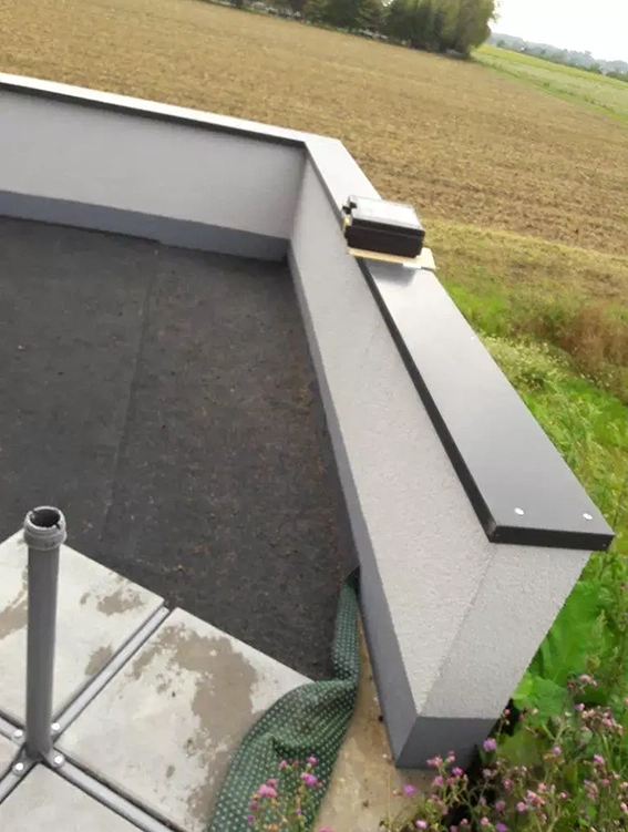 Blick von einer Dachterrasse mit grauem Bodenbelag und niedriger weißer Brüstung auf ein Feld und Wiesen im Hintergrund