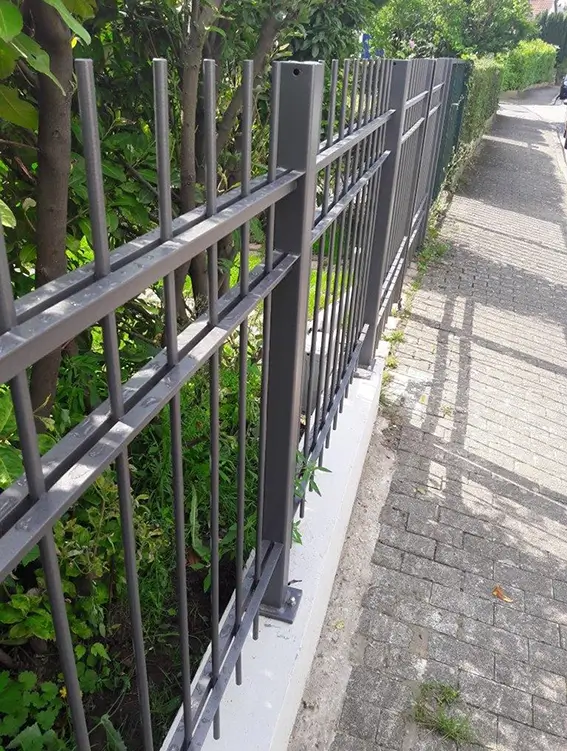 Metallzaun mit senkrechten Stäben entlang eines Gehwegs und grünem Bewuchs dahinter