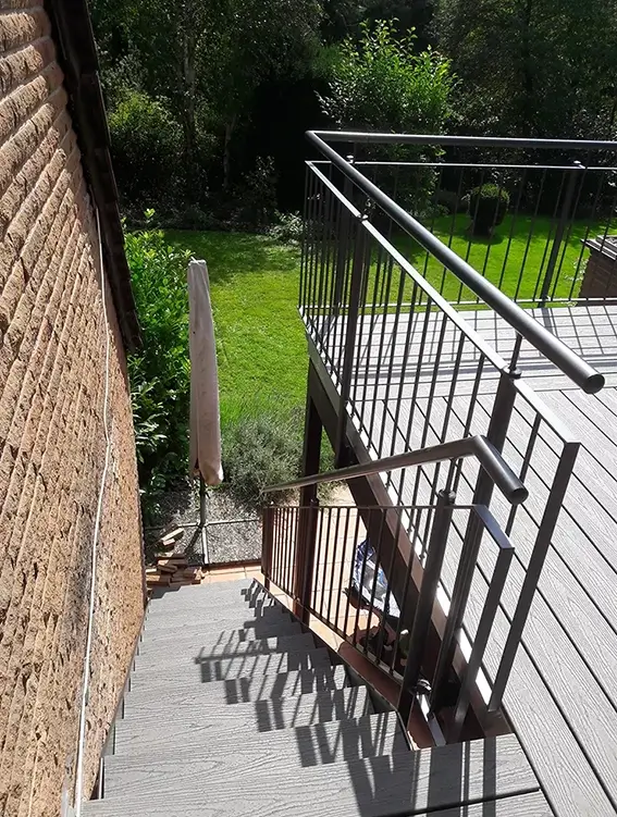 Treppe mit Metallgeländer führt von einer Terrasse aus Holz zu einem Garten mit grünem Rasen und einer geschlossenen Sonnenschirmhülle