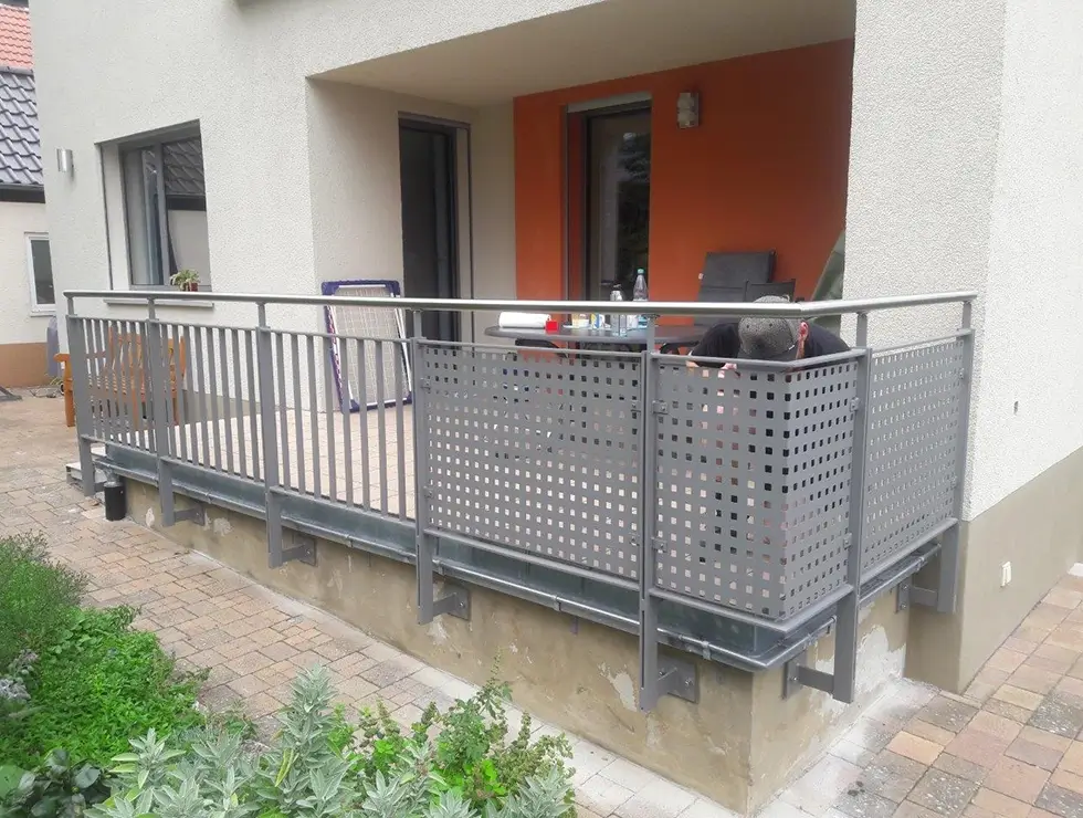 Balkon mit grauem Metallgeländer und Lochblechverkleidung an einem hellen Haus mit oranger Tür