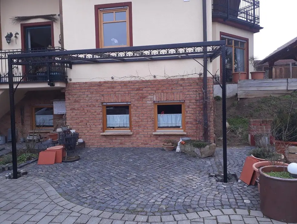 Freistehendes schwarzes Metallgestell auf gepflastertem Hof vor einem zweistöckigen Haus mit zwei Fenstern und Balkon