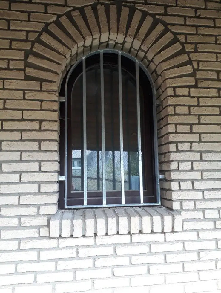 Fenster mit Rundbogen und vertikalen Metallstäben in einer hellen Ziegelmauer