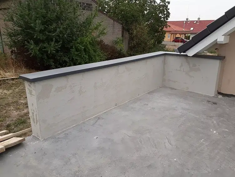 Betonterrasse mit niedriger Mauer und grauem Mauerabdeckung, im Hintergrund Bäume und ein Haus mit rotem Dach