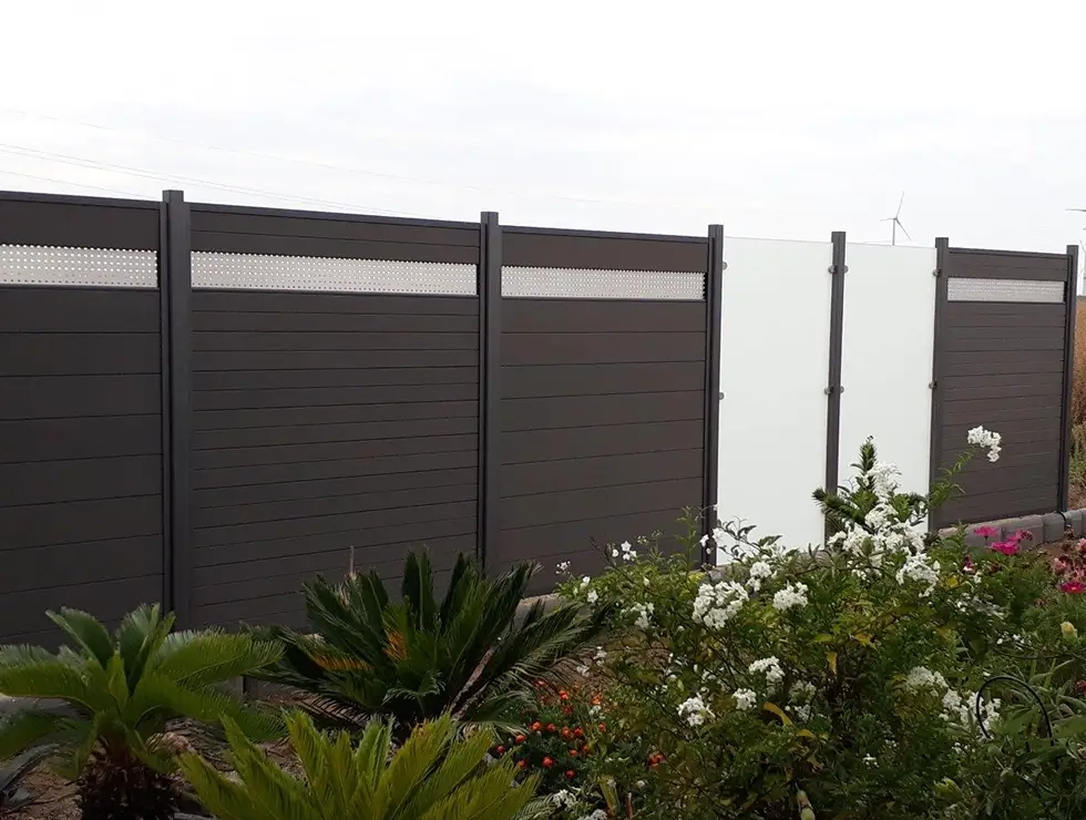Glatte, dunkelgraue Sichtschutzwand mit vertikalen Pfosten und durchbrochenem oberen Streifen vor einem Garten mit verschiedenen Pflanzen und Blumen