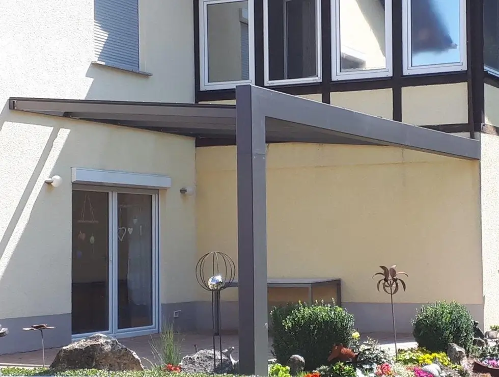 Terrassenüberdachung mit Glasdach und schwarzem Metallrahmen an einem hellen Haus mit drei Fenstern