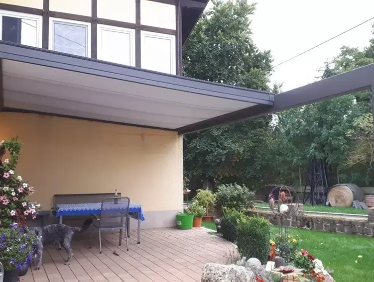 Terrasse mit Tisch, Stühlen und einem Hund unter dem Tisch neben einem Haus mit gelber Fassade und weißen Fenstern