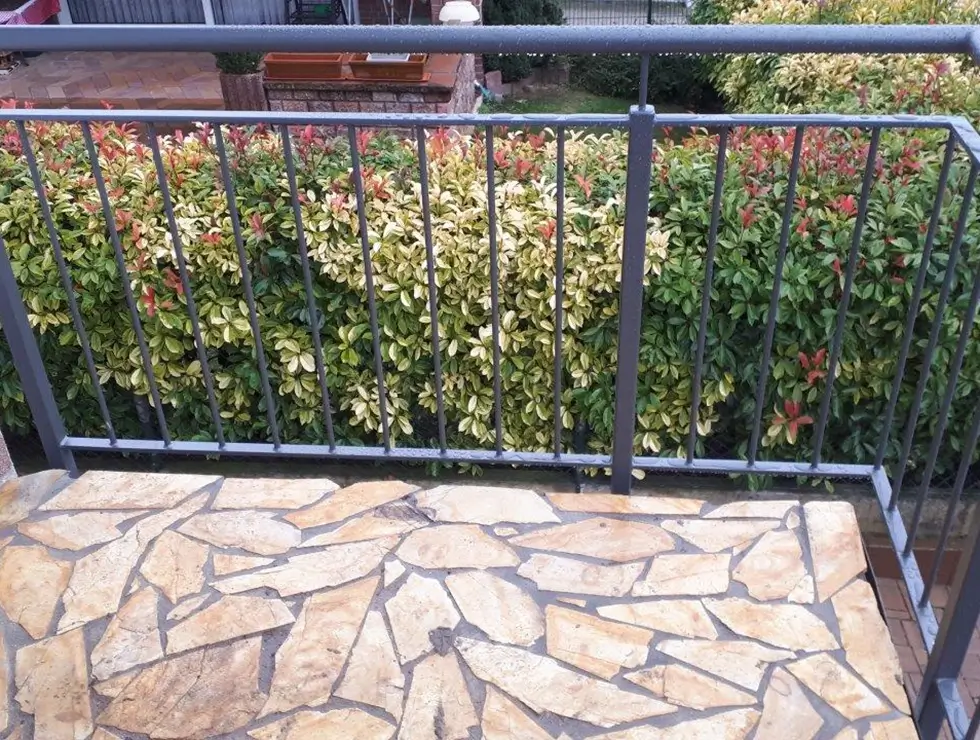 Balkon mit unregelmäßig gemustertem Steinboden und schwarzem Metallgeländer vor einer Hecke mit grünen und roten Blättern