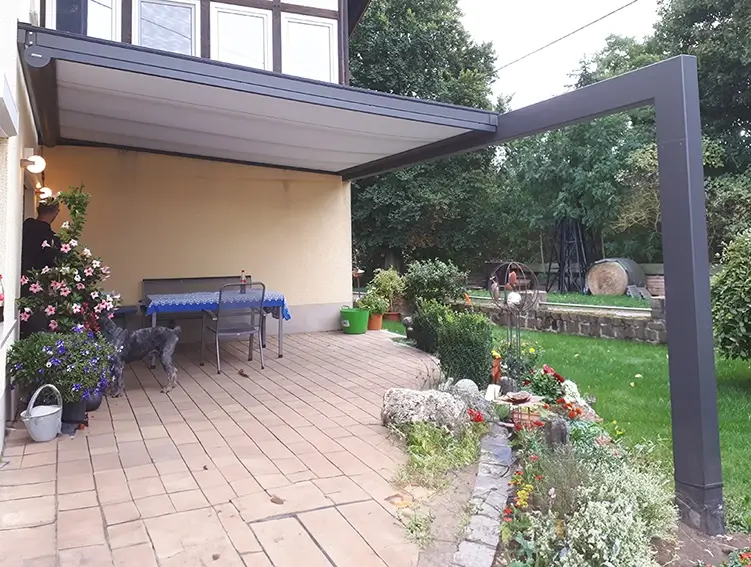 Terrasse mit Tisch und Stühlen unter einem Vordach neben einem Garten mit Blumenbeeten und Rasen