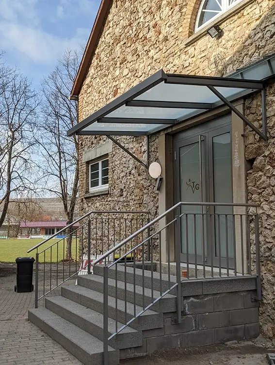 Eingang mit Treppe und Glasvordach an einem Gebäude aus Naturstein mit grauer Tür und Metallgeländer
