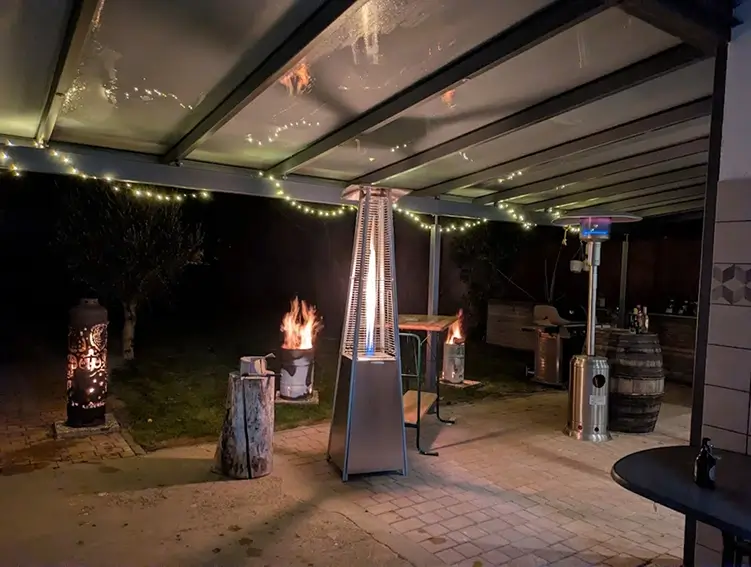 Überdachte Terrasse mit mehreren Heizstrahlern, einem Feuerkorb und Lichterkette an der Decke bei Nacht