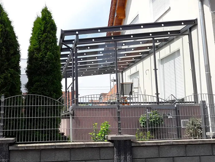 Terrasse mit Glasdach und Metallgestell neben einem weißen Haus und zwei hohen Nadelbäumen
