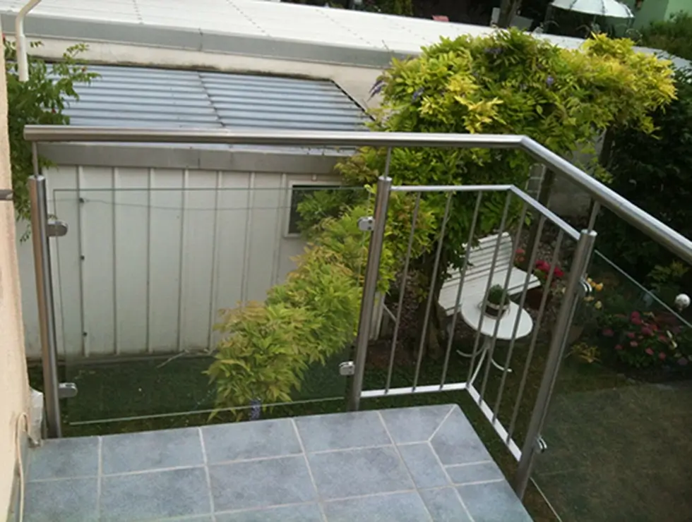 Balkon mit grauen Fliesen und Edelstahlgeländer über einem Garten mit grünem Busch und weißem Gartentisch mit Stuhl