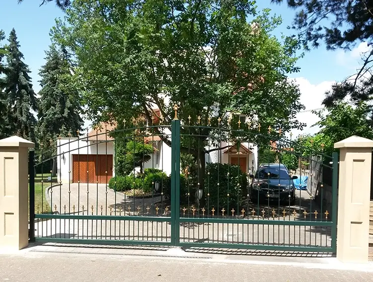 Einfahrt mit grünem Metalltor vor einem Haus mit Garage und parkendem Auto, umgeben von Bäumen
