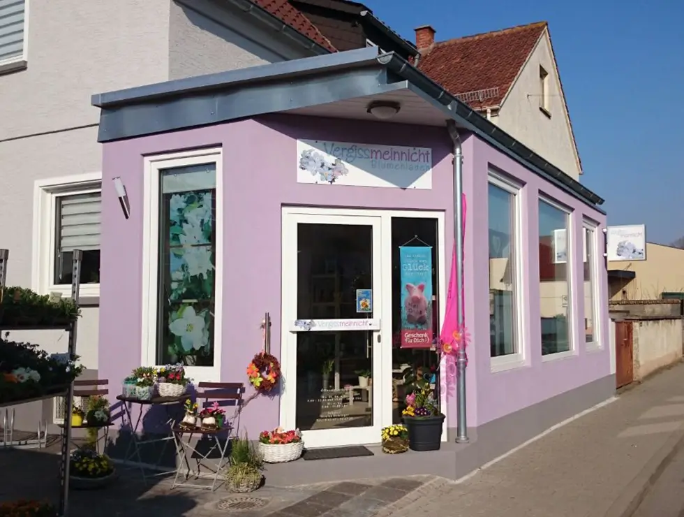 Laden mit lila Fassade, Glastür und großen Fenstern, davor Blumen und ein kleiner Tisch mit Stühlen