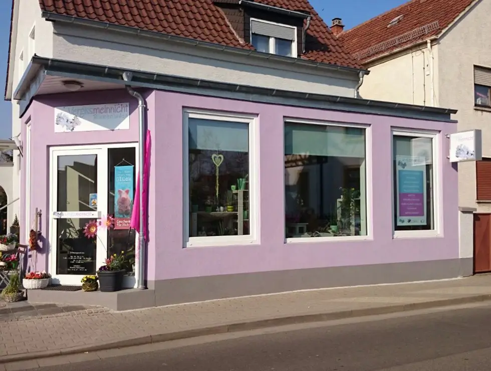 Laden mit lila Fassade, Glastür und großen Fenstern, davor Blumen und ein kleiner Tisch mit Stühlen