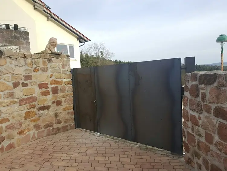 ZäuEinfahrt mit schwarzem Metalltor zwischen zwei Mauern aus unregelmäßigen Natursteinen und einer Löwenfigur auf der linken Mauer