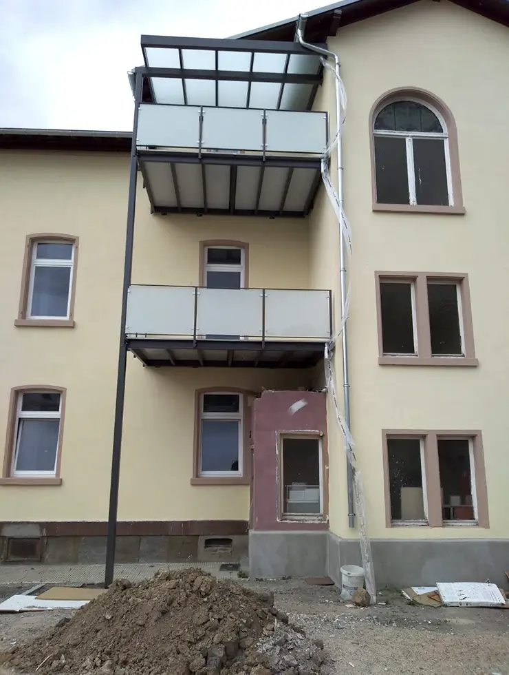 Fassade eines mehrstöckigen Hauses mit zwei Balkonen, einem kleinen Anbau und einem Erdhaufen davor