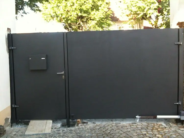 Großes, schwarzes Metalltor mit kleinem Briefkasten und Türgriff vor gepflastertem Boden und Bäumen im Hintergrund