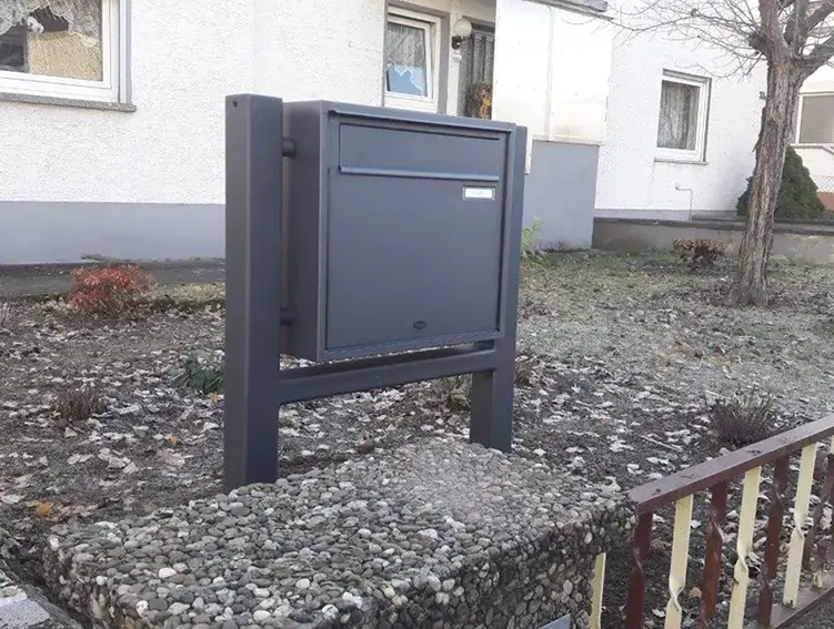 Freistehende, dunkelgraue Briefkastenanlage mit zwei Pfosten vor einem Wohnhaus mit weißer Fassade