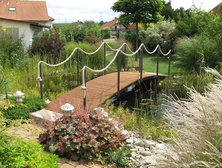 Holzbrücke mit Seilgeländer über einen kleinen Teich in einem Garten mit Rasen und Büschen