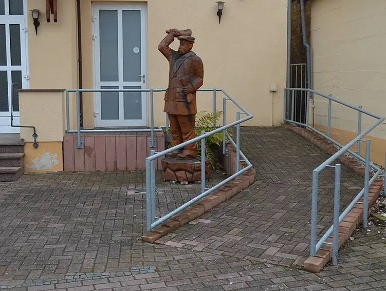 Bronzefigur eines Mannes mit Mantel und Mütze, der die Hand zum Gruß hebt, auf einem Sockel vor einem Gebäude mit Rampe und Geländer