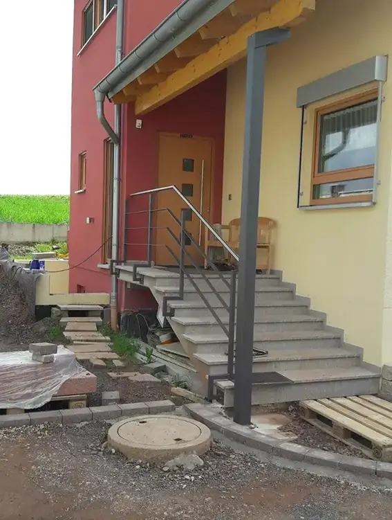 Außentreppe mit Metallgeländer und grauen Stufen an einem zweifarbigen Haus mit roter und gelber Fassade