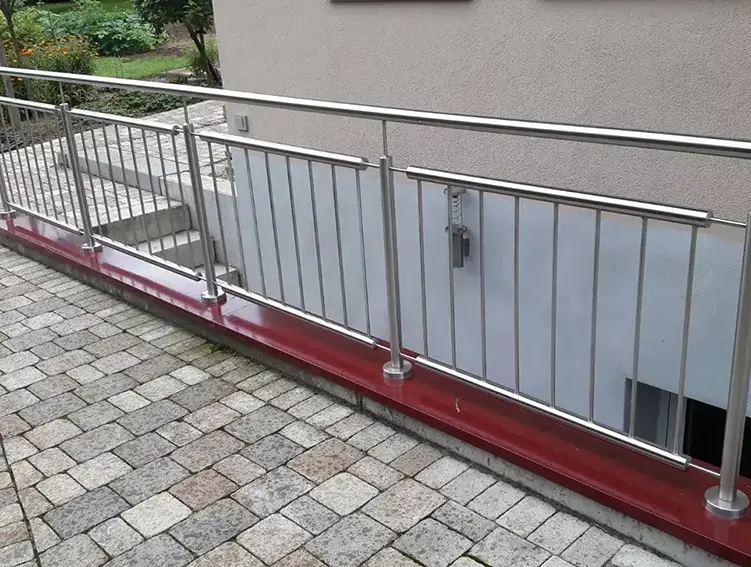 Edelstahlgeländer mit vertikalen Stäben und Handlauf an einer Treppe und Rampe neben einem Gebäude mit gepflastertem Boden