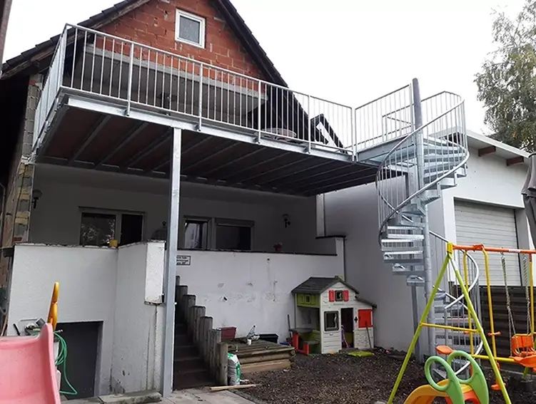 Außenbereich eines Hauses mit einer Terrasse, die über eine gerade Treppe und eine Wendeltreppe erreichbar ist, darunter Spielhaus und Kinderspielgeräte