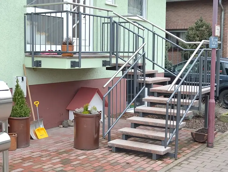 Außentreppe mit Metallgeländer und Steinstufen an einem Haus mit gepflastertem Boden und mehreren Pflanzgefäßen