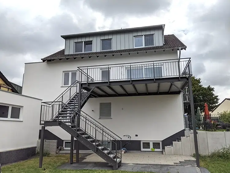 Zweistöckiges Haus mit außenliegender Metalltreppe und Balkon unter bewölktem Himmel
