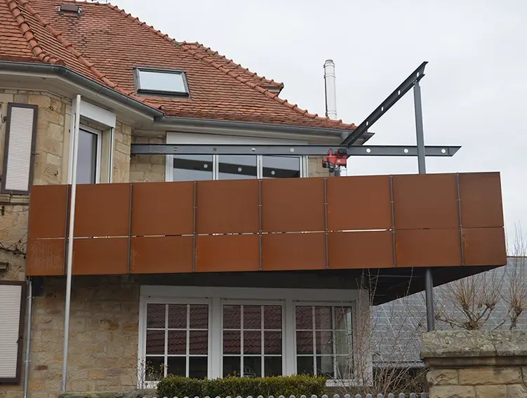 Balkon mit brauner Verkleidung an einem Haus mit rotem Ziegeldach und Steinmauer im Vordergrund