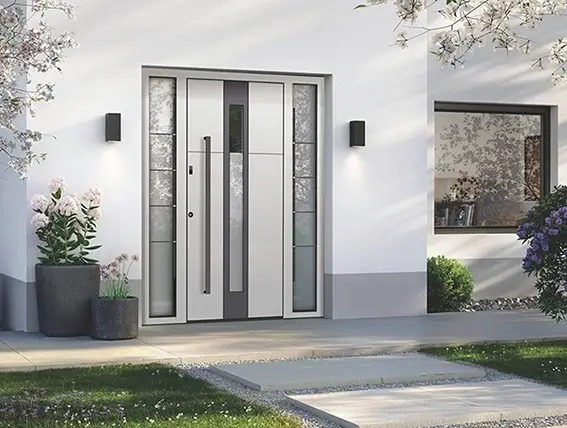 Moderner Hauseingang mit breiter Tür aus Glas und Metall, flankiert von schmalen Fenstern und zwei Wandlampen, davor Pflanzkübel mit Blumen und ein gepflasterter Weg