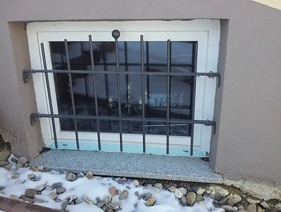 Kleines Kellerfenster mit schwarzem Gitter und Schneeresten auf dem Boden davor
